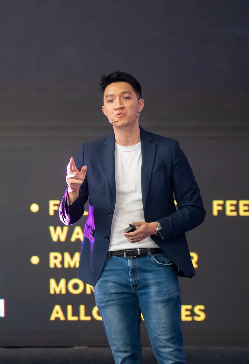 Daren Tan presenting on stage in a navy blazer and white T-shirt, gesturing as he speaks, with a slide clicker in his other hand
